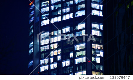 Big glowing windows in modern office building at night, in rows of windows light shines. Fragment of the glass facade of a modern corporate building at night. Modern glass office in city. Big glowing windows in modern office building at night, in rows of windows light shines. Fragment of the glass facade of a modern corporate building at night. Modern glass office in city. 135979447