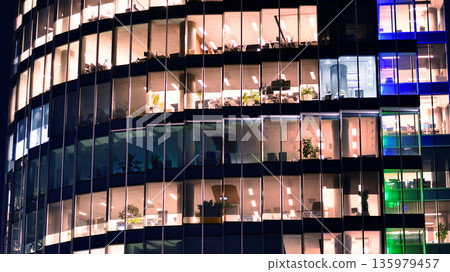 Big glowing windows in modern office building at night, in rows of windows light shines. Fragment of the glass facade of a modern corporate building at night. Modern glass office in city. Big glowing windows in modern office building at night, in rows of windows light shines. Fragment of the glass facade of a modern corporate building at night. Modern glass office in city. 135979457