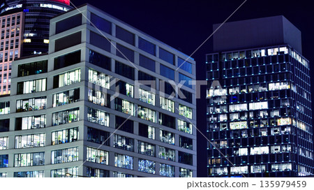 Big glowing windows in modern office building at night, in rows of windows light shines. Fragment of the glass facade of a modern corporate building at night. Modern glass office  in city. 135979459