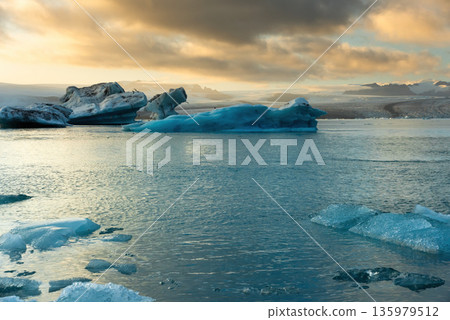 Icebergs float in Iceland's glacier lagoon, reflecting the warm colors of sunset. Birds fly above, adding life to the tranquil scene. This location is a must-see for nature enthusiasts. 135979512