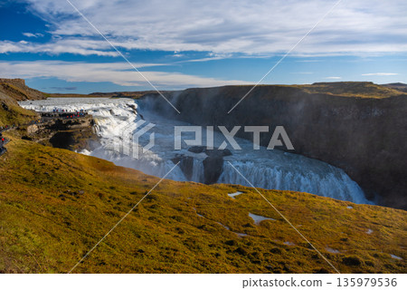 Gullfoss waterfall in Iceland showcases powerful cascades flowing into a canyon. Visitors admire the natural beauty from nearby viewpoints, especially during sunny weather. 135979536