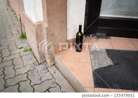 Empty blank wine bottle lying on a building entrance step. Everyday urban scene showing a discarded beverage container, representing casual street life, alcoholism, mobility, and seasonal activity. 135979931