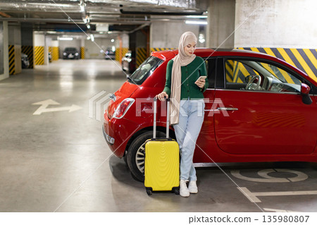 A woman wearing a hijab stands next to her car in a parking garage, looking at her phone A woman wearing a hijab stands next to her car in a parking garage, looking at her phone 135980807