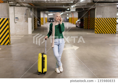 A woman wearing a hijab smiles while talking on her phone in a parking garage, holding a yellow suitcase A woman wearing a hijab smiles while talking on her phone in a parking garage, holding a yellow suitcase 135980833