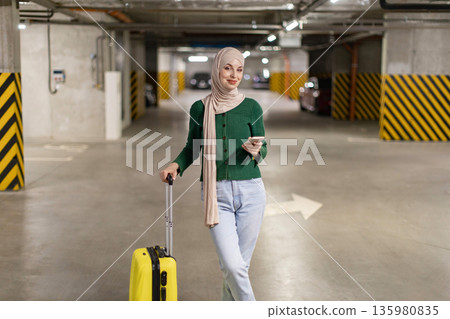 A woman wearing a hijab stands in a parking garage with her luggage, holding a phone A woman wearing a hijab stands in a parking garage with her luggage, holding a phone 135980835