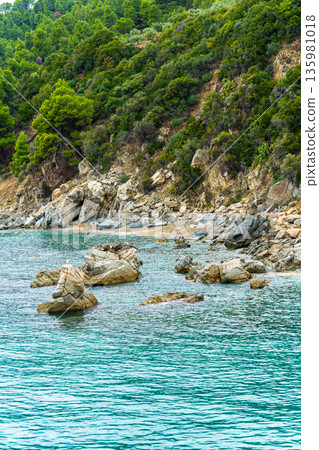 Ocean waves crash on rocks by the shore with green trees in the background near a coastal area in Greece during daylight hours Ocean waves crash on rocks by the shore with green trees in the background near a coastal area in Greece during daylight hours 135981018