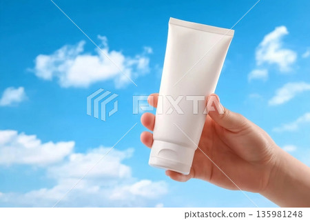 Hand of woman is holding a white plastic tube container for moisturizer or sunscreen on blue sky. Cosmetic bottle for design packaging. Minimal scene for advertising Hand of woman is holding a white plastic tube container for moisturizer or sunscreen on blue sky. Cosmetic bottle for design packaging. Minimal scene for advertising 135981248