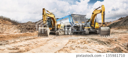 Construction activity is ongoing on dry, cracked earth where excavators are seen positioned beside a truck under a partly cloudy sky Construction activity is ongoing on dry, cracked earth where excavators are seen positioned beside a truck under a partly cloudy sky 135981710