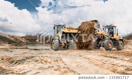 Two loaders move dirt on dry, cracked earth with dirt mounds under a partly cloudy sky during a construction activity in a large area Two loaders move dirt on dry, cracked earth with dirt mounds under a partly cloudy sky during a construction activity in a large area 135981717