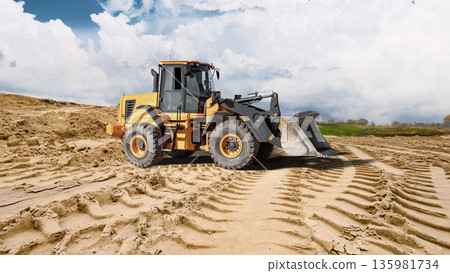 Deep tracks from heavy machinery cut through sandy ground as a loader works near a large sand pile under a cloudy sky during construction activity Deep tracks from heavy machinery cut through sandy ground as a loader works near a large sand pile under a cloudy sky during construction activity 135981734