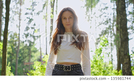 Stylish beautiful woman in casual clothing looks at the camera in warm sunset sunlight, standing in a sunlit forest, radiating natural charm. 135981744