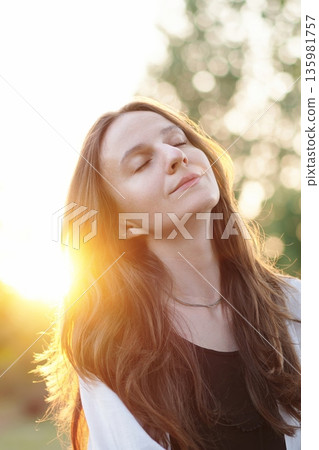 Portrait calm, beautiful young woman with closed eyes smiling at evening sunset, relieving stress after hard day in park. Serene brunette woman relaxes in the fresh air, enjoying the peace and quiet. 135981757