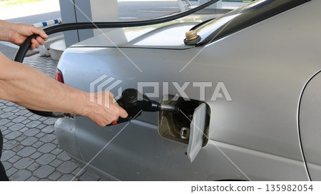 A white man's hands hold a dispensing nozzle near the open gas tank of a gray car while refueling it at a petrol station. Filling the tank with high-quality gasoline at a Russian gas station. 135982054