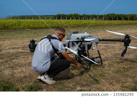 Field technician crouches beside a large agricultural drone, preparing it for field scouting and crop monitoring Field technician crouches beside a large agricultural drone, preparing it for field scouting and crop monitoring 135982295