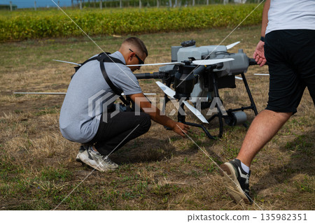 Two technicians prepare a large agricultural drone on a dry field for flight testing 135982351