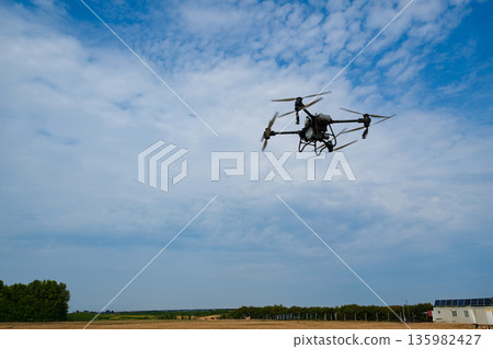 Drone in flight over open fields on a clear day with a distant farm building 135982427