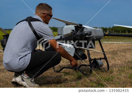 Drone operator kneels beside an agricultural drone in a dry field preparing for takeoff today Drone operator kneels beside an agricultural drone in a dry field preparing for takeoff today 135982435