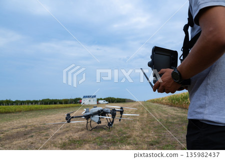 Drone pilot prepares controller while drone rests in a rural agricultural field under a blue sky Drone pilot prepares controller while drone rests in a rural agricultural field under a blue sky 135982437