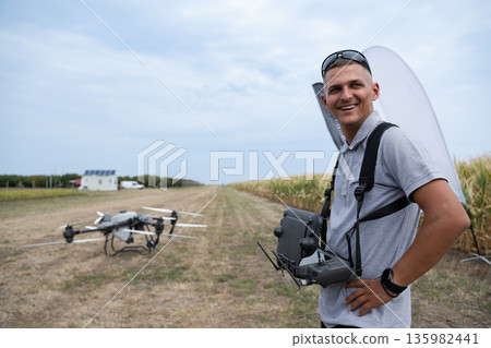Drone pilot with controller in agricultural field beside a ground drone and cornfield Drone pilot with controller in agricultural field beside a ground drone and cornfield 135982441