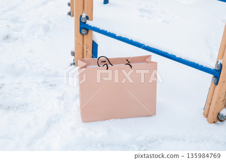 Pink paper shopping bag on white snow near a playground. Winter holiday sale concept. 135984769