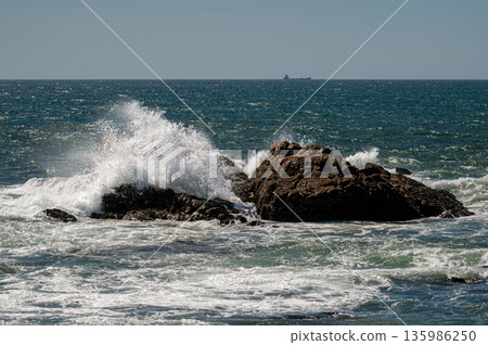 Ocean waves splashing against rocky formations while a cargo ship sails in the distance. 135986250