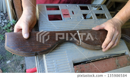 Hands of a carpenter holding a prefabricated wooden piece with curved lines and varnished, cut into two parts, top view, showing the sawn wooden turned part of the furniture on the iron work surface 135986584