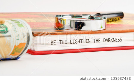 A magnifying glass rests on an open book displaying the message BE THE LIGHT IN THE DARKNESS A magnifying glass rests on an open book displaying the message BE THE LIGHT IN THE DARKNESS 135987488