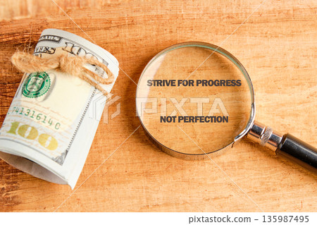 A magnifying glass focuses on the text Strive for progress not perfection on a wooden background. Beside it rests a tightly rolled bundle of one hundred dollar bills secured with twine A magnifying glass focuses on the text Strive for progress not perfection on a wooden background. Beside it rests a tightly rolled bundle of one hundred dollar bills secured with twine 135987495