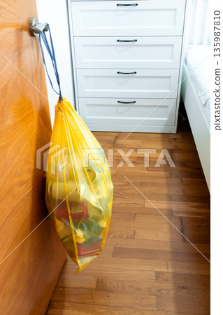 Yellow trash bag hanging on door handle in bright room with wooden floor and white furniture 135987810