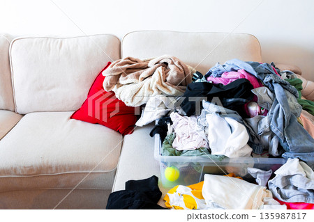 Messy laundry overflowing on beige couch and red pillow in bright living room Messy laundry overflowing on beige couch and red pillow in bright living room 135987817