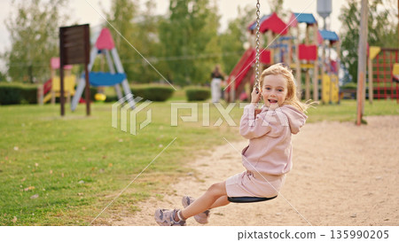 Excited girl enjoys swinging on swing. Happy child having fun on a playground, smiling brightly and experiencing joy during a playful outdoor moment. Excited girl enjoys swinging on swing. Happy child having fun on a playground, smiling brightly and experiencing joy during a playful outdoor moment. 135990205