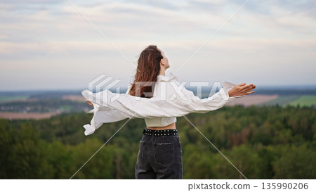 Young woman standing with raised arms on a hilltop, feeling the wind with her hair and shirt flowing. Young woman standing with raised arms on a hilltop, feeling the wind with her hair and shirt flowing. 135990206
