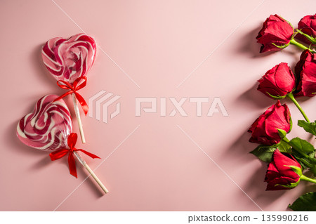 Valentine's Day background. Red roses and heart shaped lolly candy over pink background. Abstract flat lay. Celebrate, festive greeting card. Valentine's Day background. Red roses and heart shaped lolly candy over pink background. Abstract flat lay. Celebrate, festive greeting card. 135990216