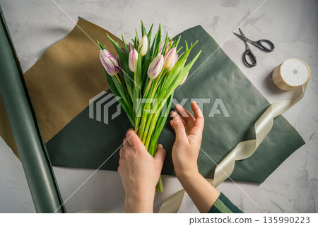 Mother's day, birthday bouquet. Female hands wrapping tulip flowers in craft paper. Spring tulips flower background Mother's day, birthday bouquet. Female hands wrapping tulip flowers in craft paper. Spring tulips flower background 135990223