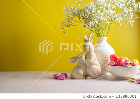 Easter chocolate colourful eggs in a bowl, cute bunnies and gypsophila flowers in a vase. Spring holiday background. 135990301