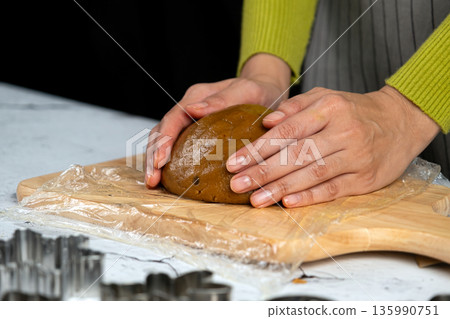 Close-up of hands kneading cookie dough on a wooden cutting board with plastic wrap, preparing homemade baked goods in a cozy kitchen Close-up of hands kneading cookie dough on a wooden cutting board with plastic wrap, preparing homemade baked goods in a cozy kitchen 135990751
