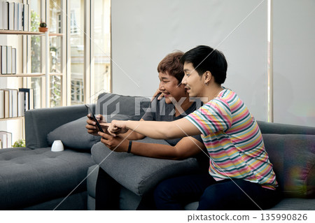 Two Asian men sitting on sofa smiling and pointing at tablet screen in cozy home. This candid moment reflects digital connection, friendship, and modern tech lifestyle Two Asian men sitting on sofa smiling and pointing at tablet screen in cozy home. This candid moment reflects digital connection, friendship, and modern tech lifestyle 135990826