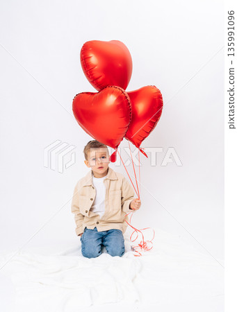Little boy sitting with red heart balloons on white background. Valentines Day, romantic holiday, childhood innocence, love and joy Little boy sitting with red heart balloons on white background. Valentines Day, romantic holiday, childhood innocence, love and joy 135991096