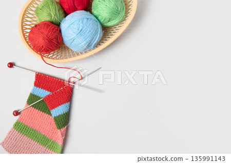 Red, green, blue and pink knitting balls in basket with knitting needles and knitted scarf on gray background. Set for the hobby of knitting warm clothes or toys. Concept needlework Red, green, blue and pink knitting balls in basket with knitting needles and knitted scarf on gray background. Set for the hobby of knitting warm clothes or toys. Concept needlework 135991143