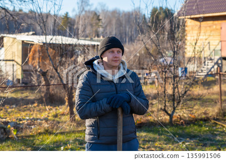 Young man standing with shovel in yard. Gardening and land work concept. Rural spring or autumn outdoor activity. Young man standing with shovel in yard. Gardening and land work concept. Rural spring or autumn outdoor activity. 135991506