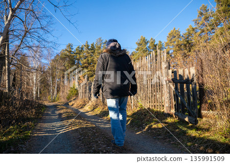 Man walks along a dirt road in a village with a wooden fence and trees on a sunny day. Rural nature and outdoor adventure. Man walks along a dirt road in a village with a wooden fence and trees on a sunny day. Rural nature and outdoor adventure. 135991509
