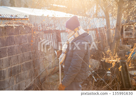 man sweeps yard in sun's rays. Man holding broom wearing winter clothes, surrounded by dry vegetation. Spring cleaning or yard work on a sunny day. man sweeps yard in sun's rays. Man holding broom wearing winter clothes, surrounded by dry vegetation. Spring cleaning or yard work on a sunny day. 135991630
