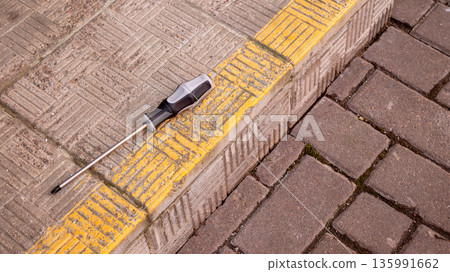 Screwdriver with magnetic tip on a yellow tactile paving on a sidewalk. Tool for repair and construction. 135991662