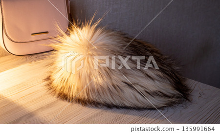 Fluffy fur hat or pompom accessory on a wooden table. Warm winter headwear for cold weather fashion and cozy style. Fox fur on a knitted base 135991664