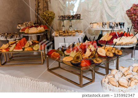 Elegant Fruit and Dessert Buffet Display with Assorted Sliced Fruits, Pastries, and Decorative Glass Stands in Luxury Event Setting Elegant Fruit and Dessert Buffet Display with Assorted Sliced Fruits, Pastries, and Decorative Glass Stands in Luxury Event Setting 135992347