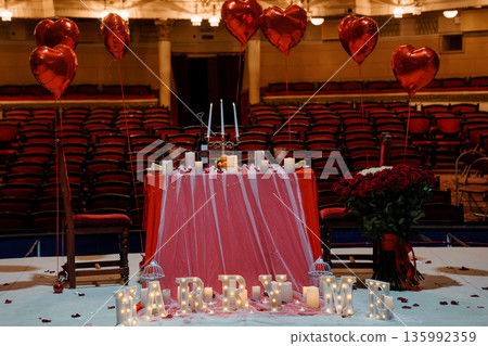 Romantic Proposal Setup with Heart Balloons, Rose Bouquet, and "MARRY ME" Lights in a Grand Theater 135992359