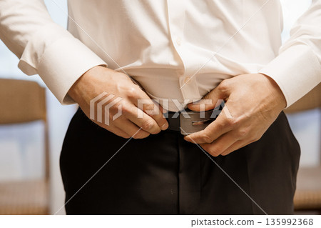 Man in White Dress Shirt Adjusting Black Belt While Wearing Formal Black Trousers in Bright Indoor Setting 135992368