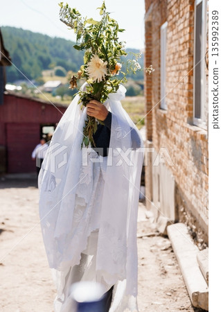 Bride in White Veil Holding Flower Bouquet in Rural Village Street with Brick House and Green Hills Background Bride in White Veil Holding Flower Bouquet in Rural Village Street with Brick House and Green Hills Background 135992389