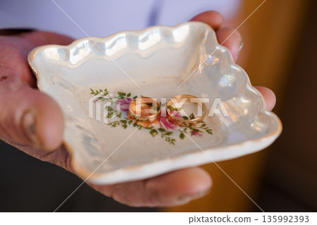 Elegant Gold Wedding Rings Resting in a Vintage Porcelain Dish with Floral Design Held by Hand 135992393