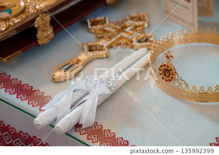 Golden Orthodox Christian wedding cross and crown with white candles on embroidered tablecloth, religious ceremony symbols Golden Orthodox Christian wedding cross and crown with white candles on embroidered tablecloth, religious ceremony symbols 135992399